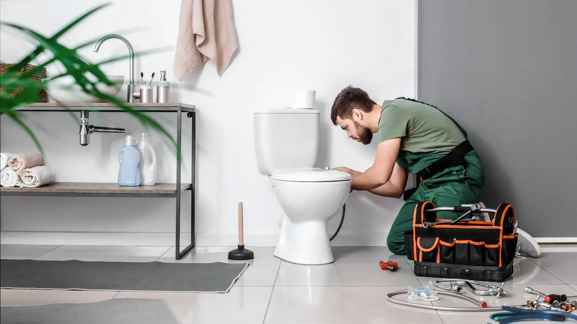 Plumber in green overalls repairing a white toilet in a modern bathroom. Tools and a toolbox are nearby.