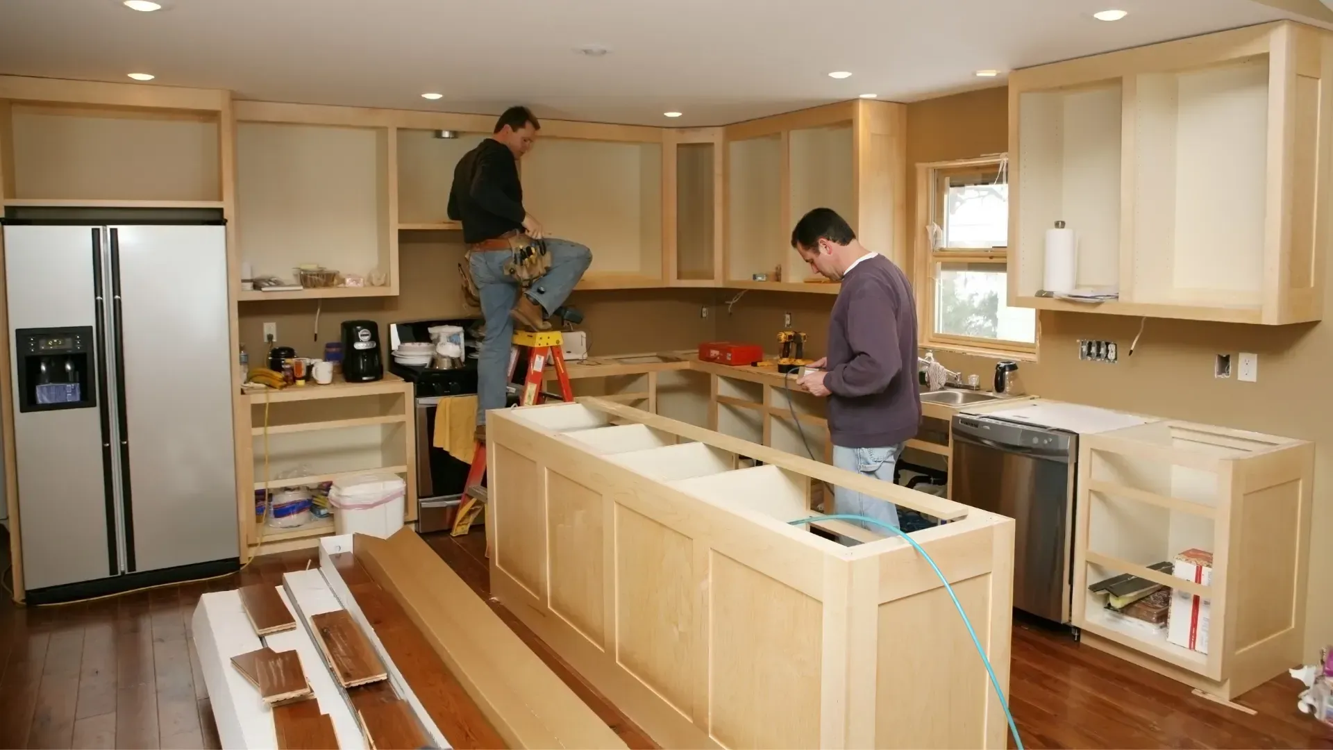Two men installing wooden kitchen cabinets, island in the foreground, appliances and tools are visible.