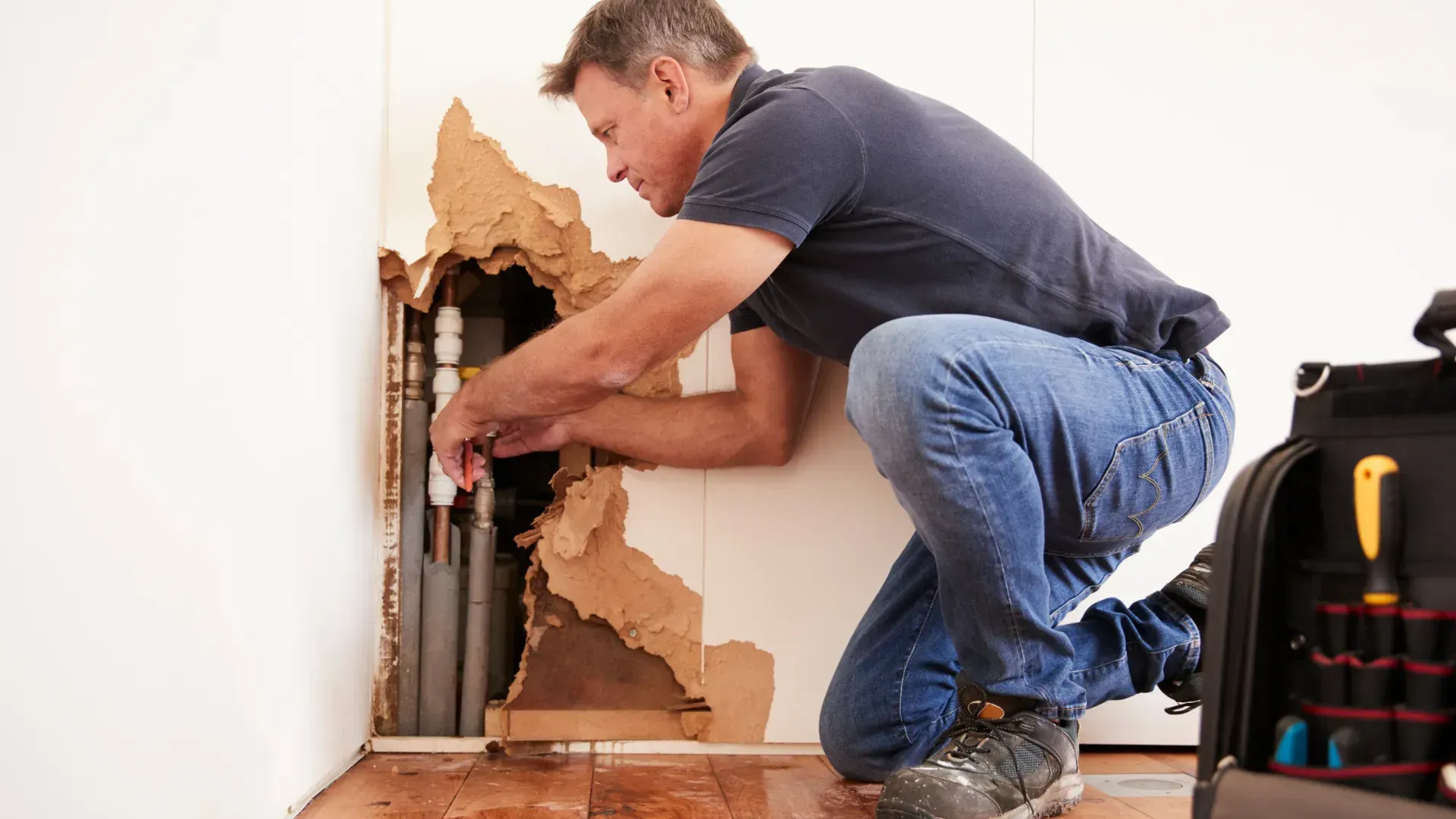 Plumber kneeling to repair pipes in a wall, with damaged drywall. He's wearing jeans and a blue shirt.