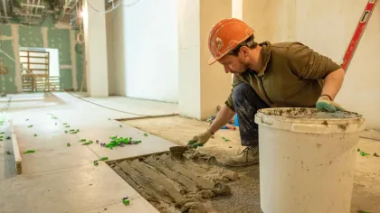 A construction worker, wearing a hard hat, spreads mortar with a trowel on a floor.