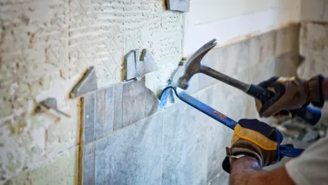 Person removing bathroom tile with a hammer and pry bar, wearing gloves in a dusty setting.
