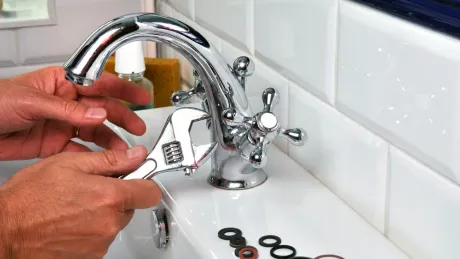 Hands using a wrench to repair a chrome bathroom faucet; several washers are in the sink.