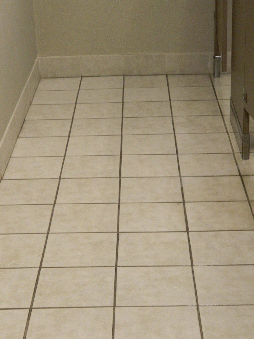 Tiled bathroom floor, beige tiles with dark grout, leading towards a stall.