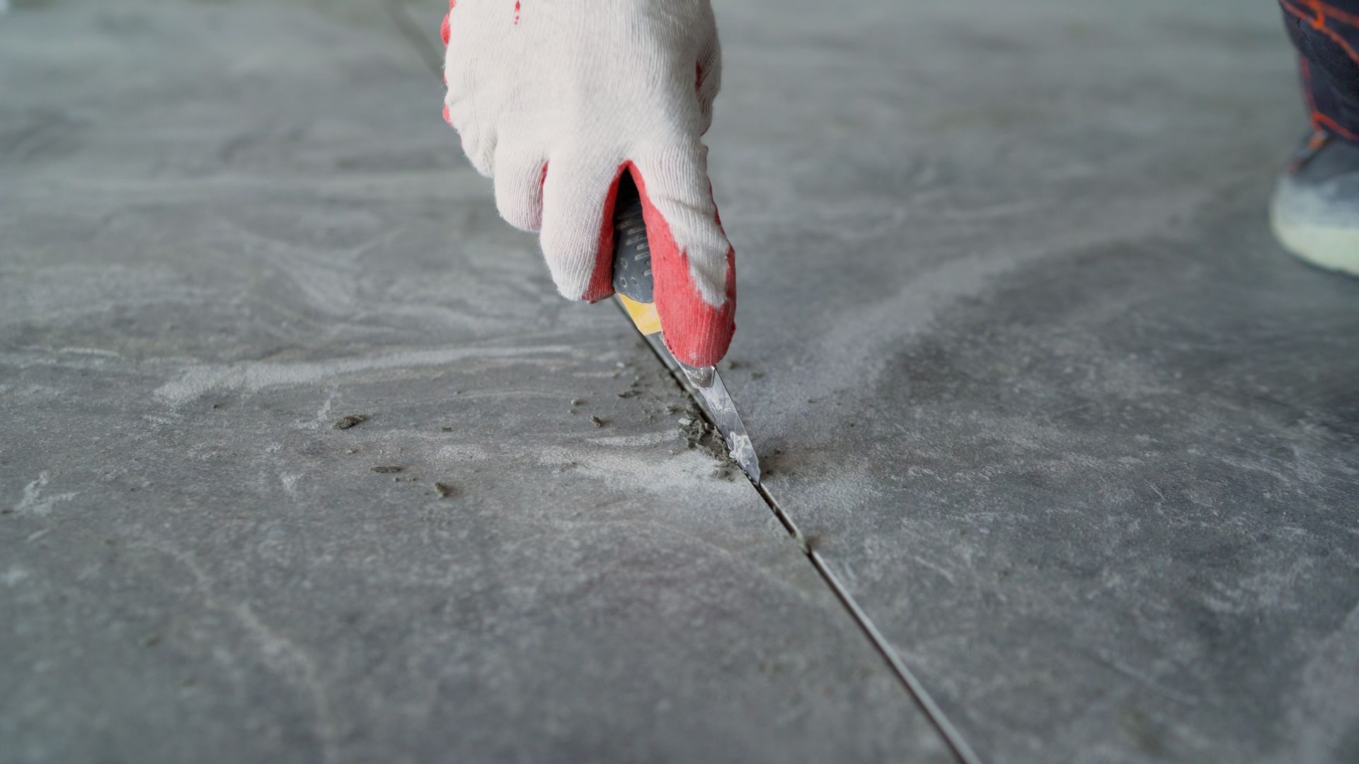 Gloved hand using a utility knife to cut into a gray concrete floor's grout line, indoor setting.