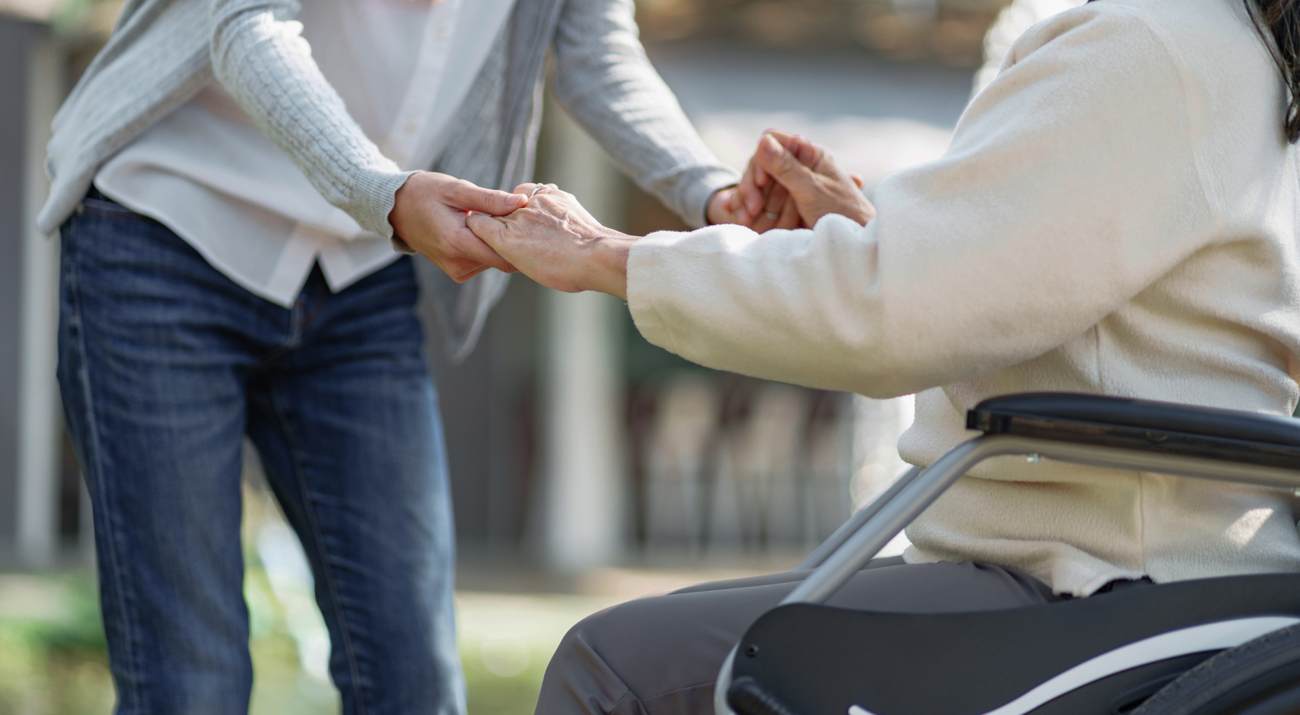 Woman in wheelchair holds hands with a person in jeans and a gray sweater outdoors.