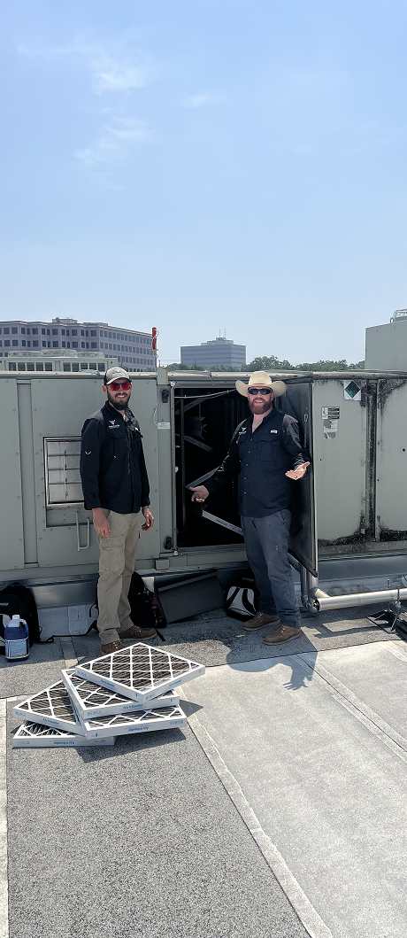 Two men on a rooftop by HVAC equipment, one pointing inside. Filters on ground, blue sky.