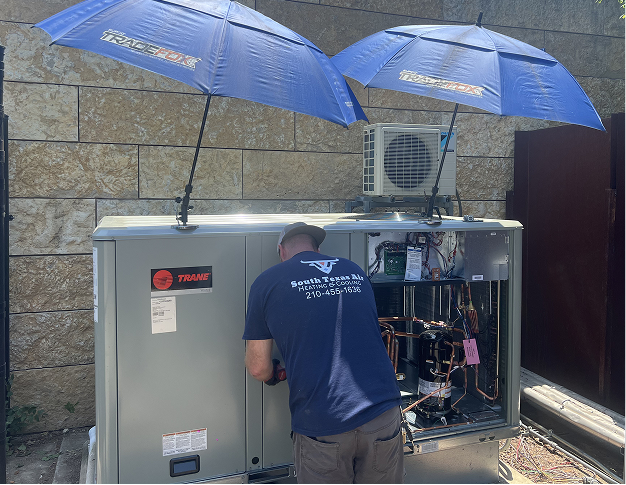 HVAC technician working on an air conditioning unit under two blue umbrellas, outdoors.