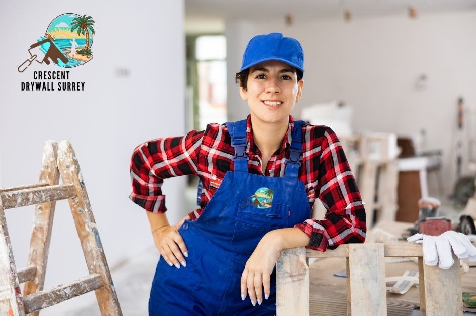 A drywall repair company worker in blue overalls, red plaid shirt, and blue cap smiles while leaning against a stepladder in a residential renovation space with white walls and construction materials visible near Fortress Dr, and Cannon Green in Port Coquitlam, BC.