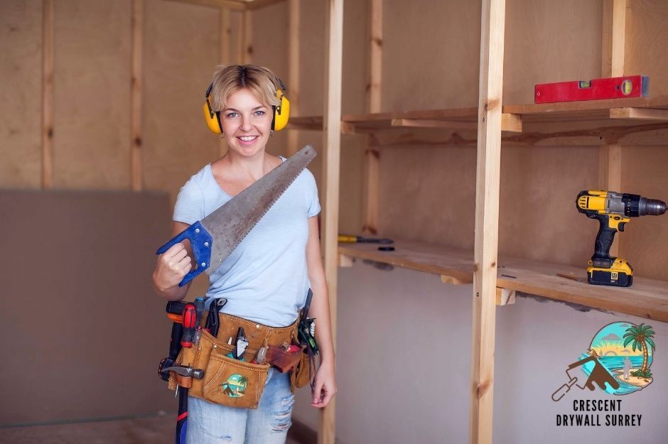 A drywall repair company worker wearing yellow ear protection and a tool belt smiles while holding a saw in a residential construction space with exposed wooden framing and tools visible near Buena Vista Ave, and Vidal St in White Rock, BC.