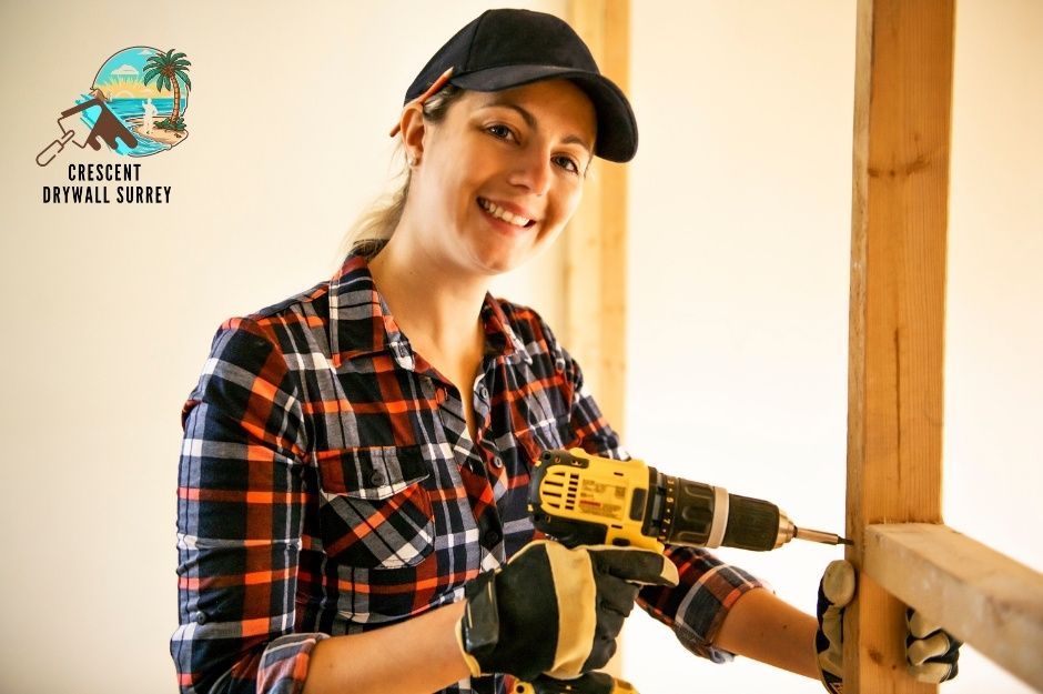 A smiling female drywall repair contractor wearing a plaid shirt and a baseball cap holds a yellow cordless drill while working on wooden framing in a residential construction project. She's positioned near exposed wood studs in an unfinished interior space near 240a St, and Hill Ave in Maple Ridge, BC.