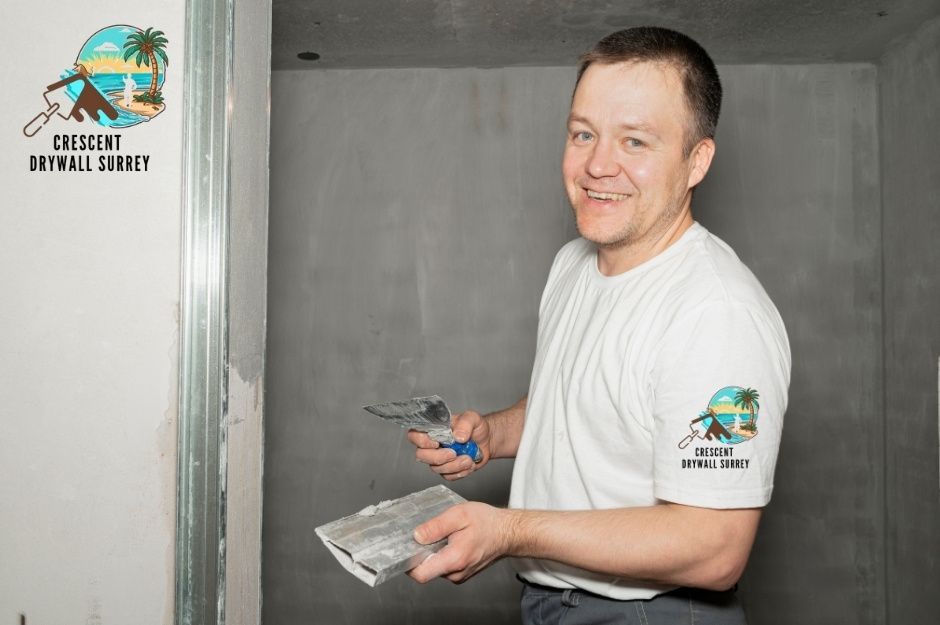 A drywall repair contractor applies joint compound with a trowel. A worker in a white shirt smiles while holding putty knife and mud pan, smoothing drywall seams on a residential interior wall with metal corner bead visible at Ioco Rd, and 1st Ave in Port Moody, BC.