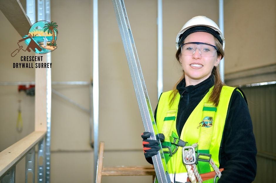 A drywall repair contractor wearing a yellow high-visibility safety vest and white hard hat smiles while holding measuring tools at a commercial construction site with metal framing and drywall panels visible near 84 Ave, and 201A St in Willoughby, BC.