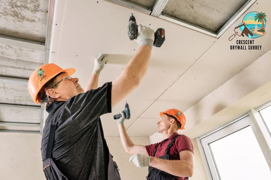 Drywall repair contractors install ceiling panels in a commercial building. Two workers wearing orange hard hats use drills to secure white drywall sheets to a ceiling framework at 10th St E, and St Andrews Ave in North Vancouver, BC.