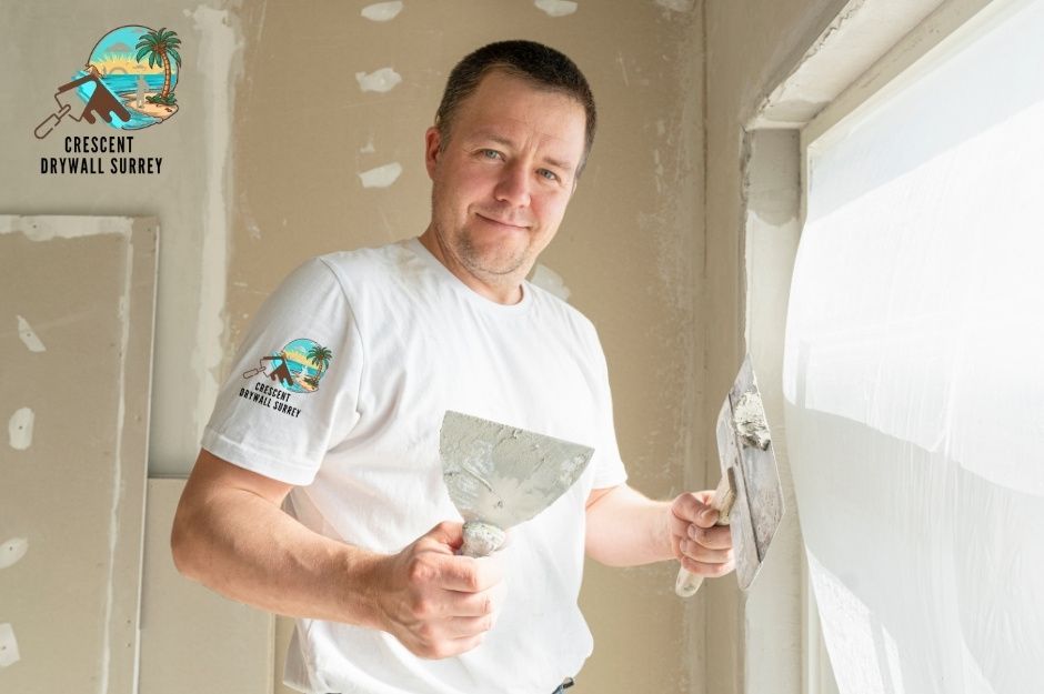 A drywall repair handyman applies joint compound to wall seams. He is in a white shirt holding a putty knife and spreader tool while smoothing drywall mud over taped joints in a residential interior room with white walls and ceiling near Sperling Ave, and Napier St in Burnaby, BC. 