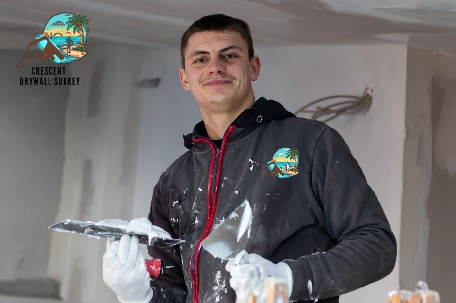 A drywall repair handyman in a dark hoodie smiles while holding a putty knife in a residential interior with white walls and ceiling fixtures visible during renovation work near Thorburn Wy, and Bonson Rd in Pitt Meadows, BC.
