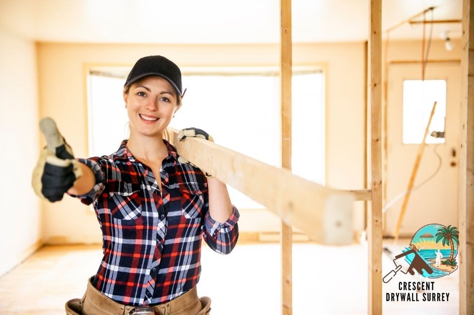 A drywall repair specialist in a plaid shirt and black cap smiles while holding wood in a residential construction space with wooden framing and bright interior lighting visible near 16 Ave, and 57 St in Delta, BC.