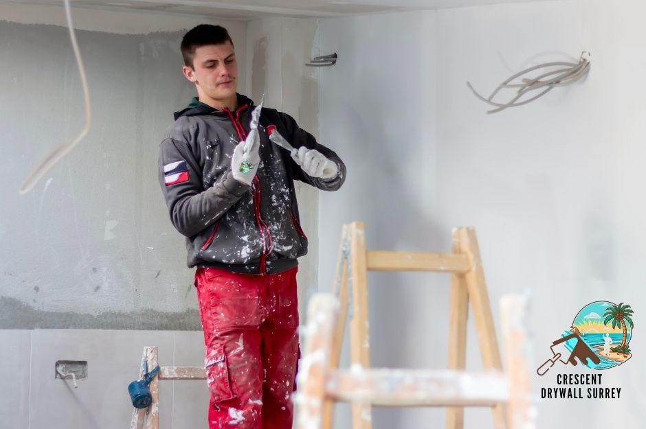 A drywall repair technician in red work pants and grey jacket holds a trowel while working on white walls in a commercial space, with a wooden ladder and electrical outlets visible near Grade Crescent, and 48 Ave in Langley, BC.