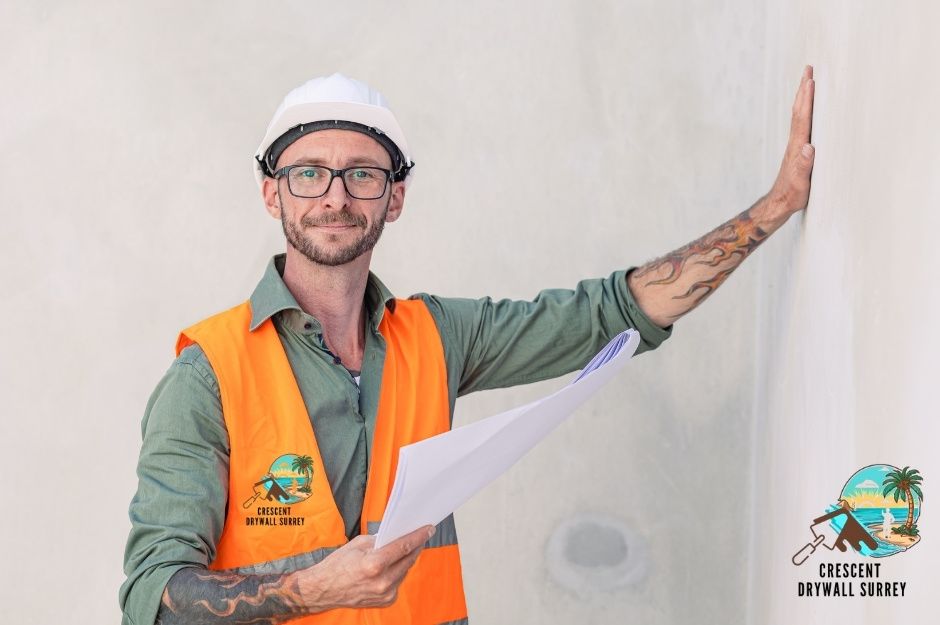 A drywall repair technician inspects the wall surface quality. The contractor is wearing an orange safety vest, glasses, and a white hard hat, extending his hand to check the smoothness of the drywall finish in a commercial building interior during a construction project at Kelly St, and Major St in New Westminster, BC.