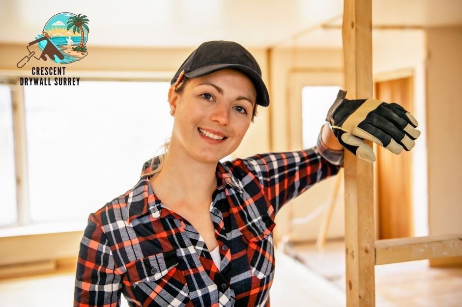 A drywall repair technician in a plaid shirt and black cap smiles while leaning against wooden framing in a commercial construction space with bright lighting and structural beams visible near Victoria Dr, and Jasper Cres in South Vancouver, BC.