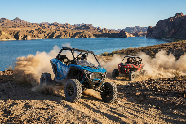 Two off-road vehicles kick up dust while driving near a lake and mountains.