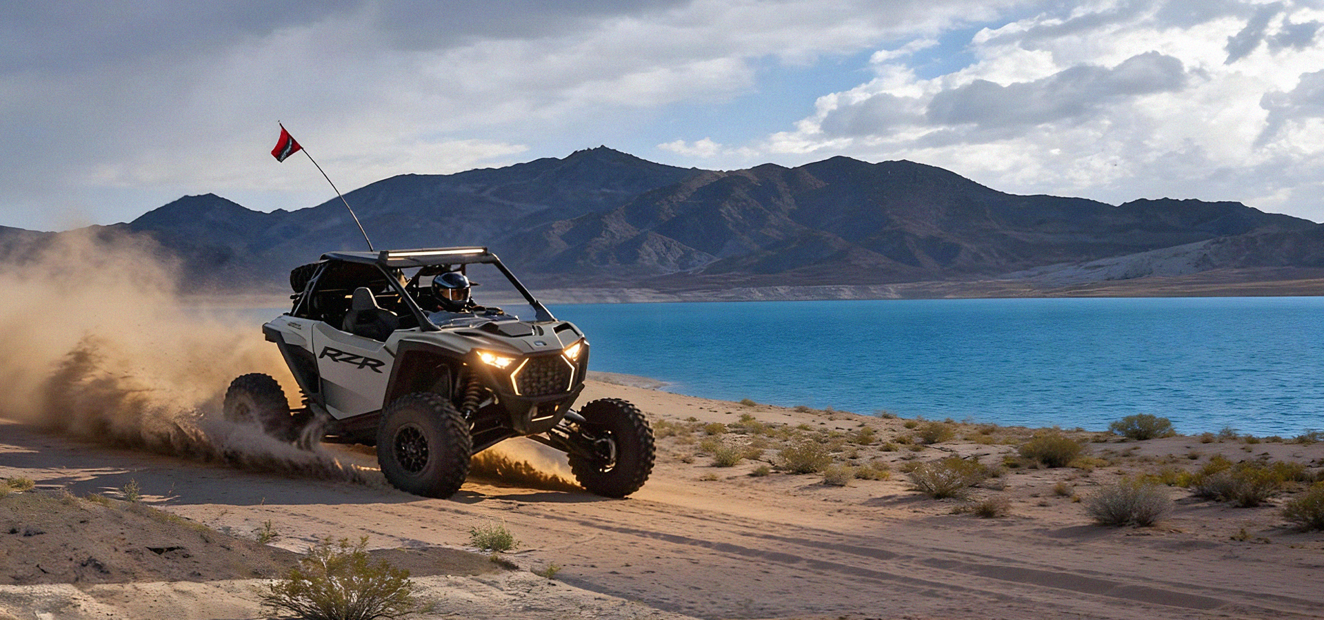 White off-road vehicle kicking up dust on a dirt road by a blue lake with mountains in the background.