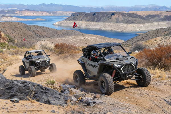 Two gray off-road vehicles kick up dust on a dirt trail, with a lake and mountains in the background.