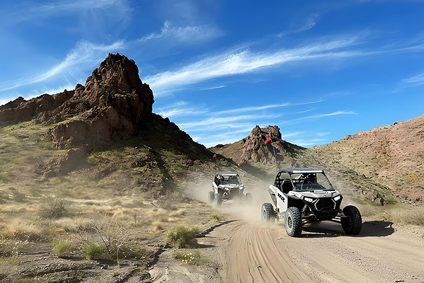 Two off-road vehicles kick up dust on a desert trail, mountains and blue sky in the background.