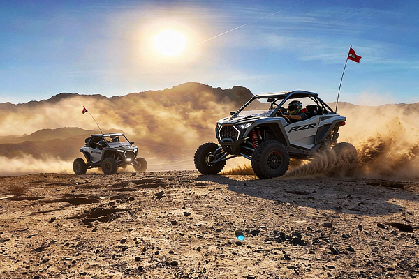 Two off-road vehicles racing across a desert terrain, kicking up dust under a bright sun.