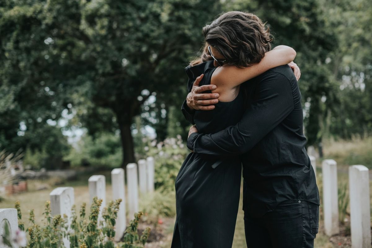 Two people hugging in a cemetery, surrounded by white gravestones and trees