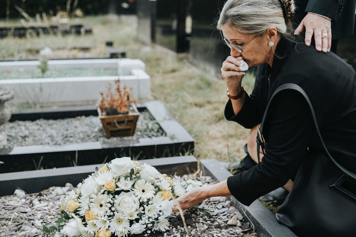 Person mourning at a graveside, placing flowers on a grave and covering their face