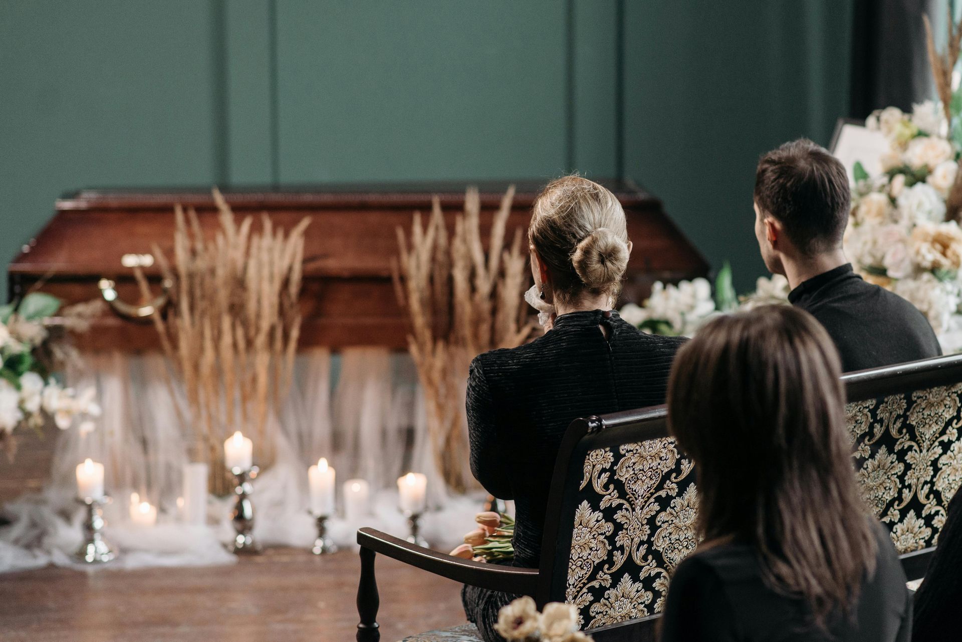 Guests seated at a candlelit funeral service facing a flower-covered casket in a church