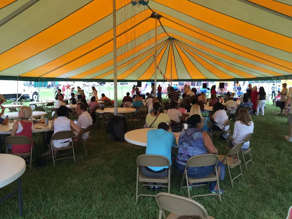 People seated at tables inside a large orange-striped tent at an outdoor gathering.