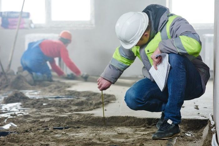 Construction worker in a hard hat inspecting flooring with a measuring tool; another worker in background.