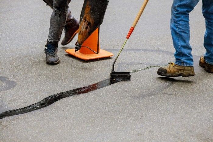 Workers sealing a crack in asphalt with a sealant and a roller, near an orange traffic cone.