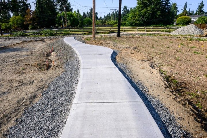 Concrete sidewalk curves through a grassy area, bordered by gravel on either side.