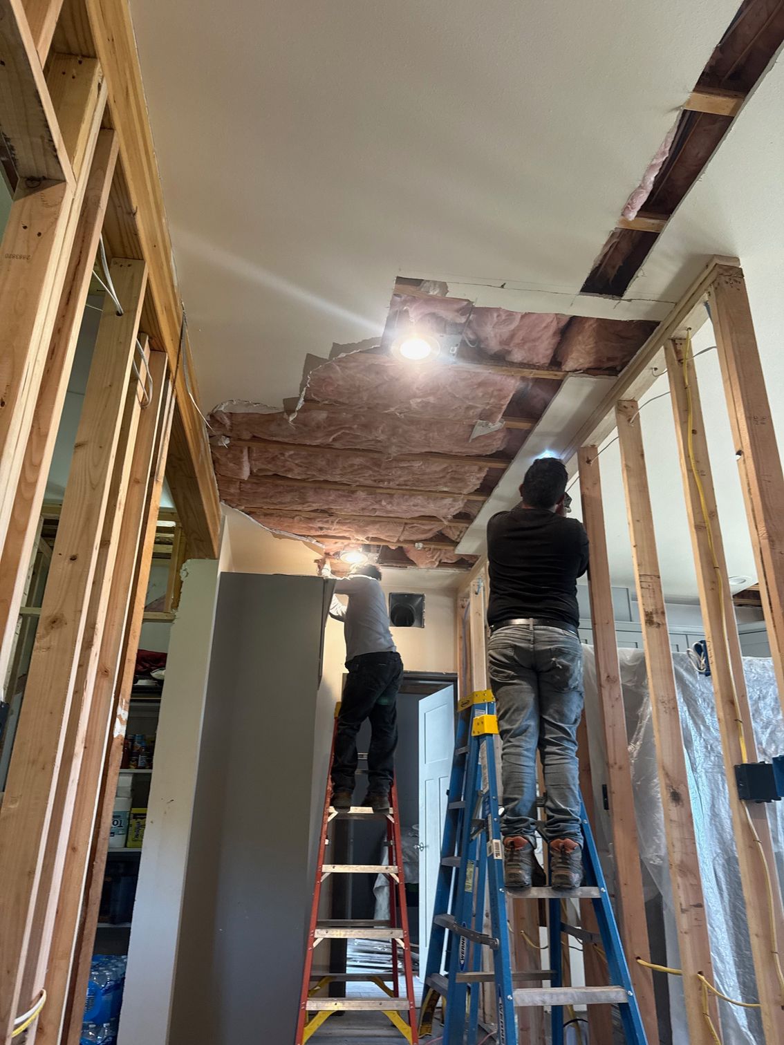 Two people on ladders removing ceiling, exposed insulation, wood framing.