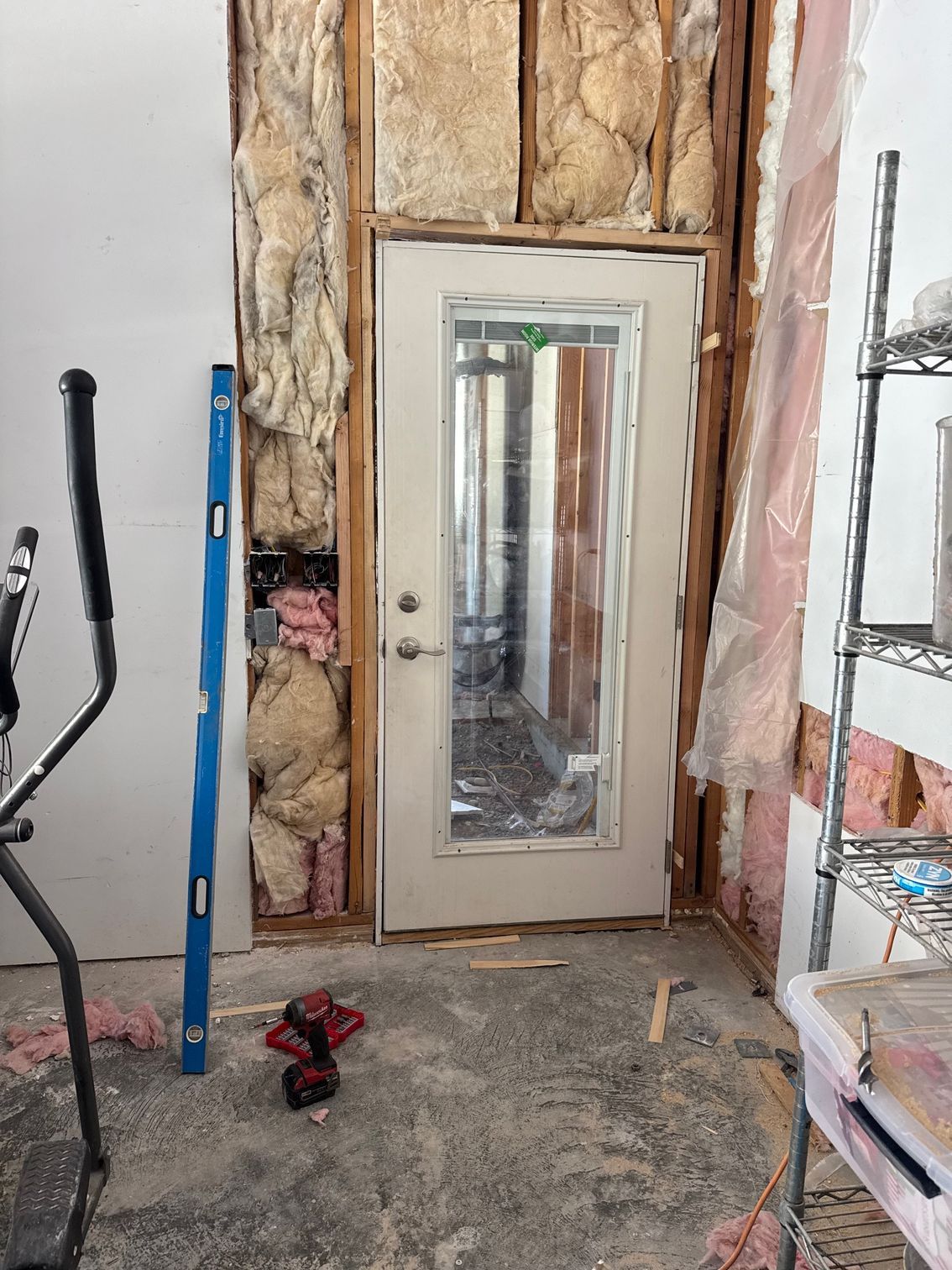 Door in partially demolished wall, insulation exposed. Blue level propped against wall; exercise equipment in foreground.