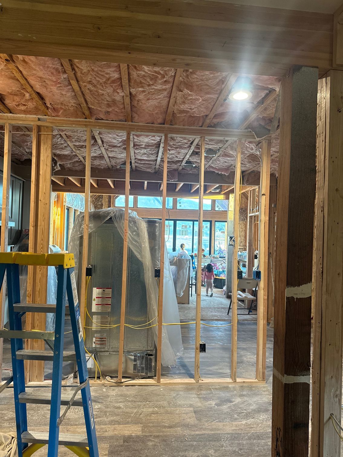 Interior construction scene: unfinished wooden framing, insulation, ladder, and a water tank covered in plastic.