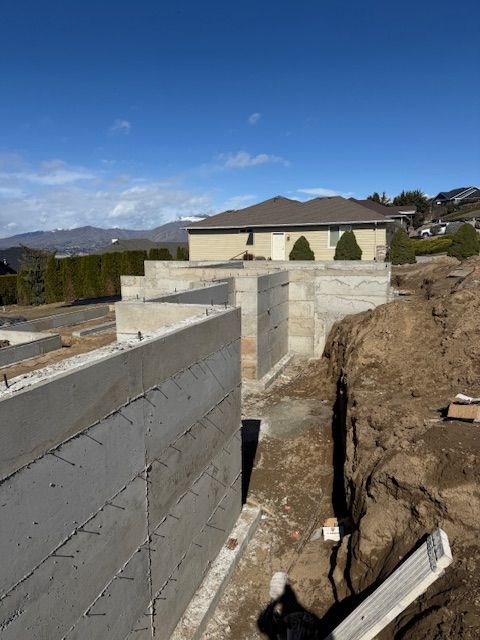 Concrete foundation walls under construction, with a house in the background against a blue sky and mountains.