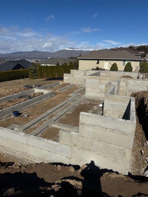 Concrete foundation of a house under construction, with surrounding dirt and a house in the background.