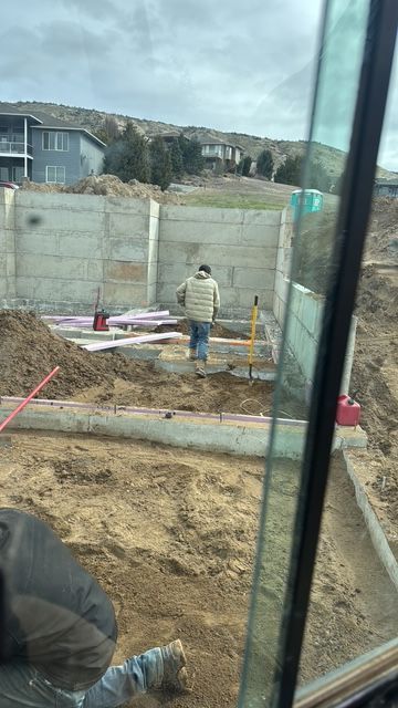 Construction worker inside foundation, digging with a shovel. Gray concrete walls, brown dirt.