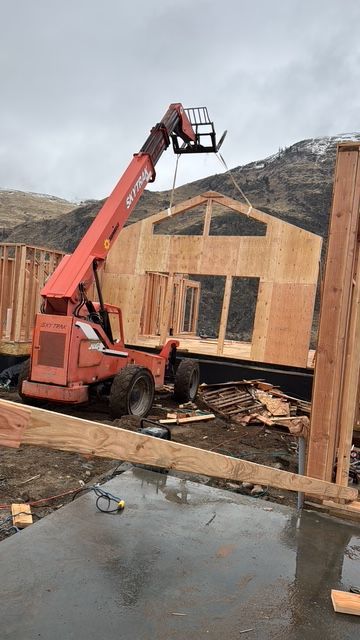 A red forklift lifting a wooden wall panel for a house under construction; mountains in background.