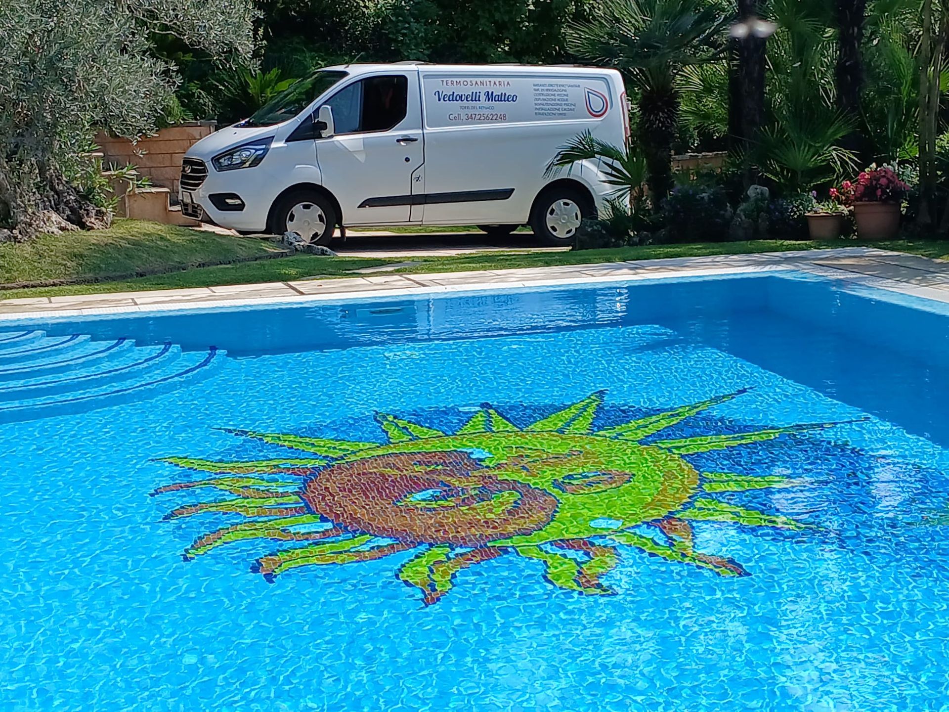 Furgone bianco parcheggiato vicino a una piscina con un mosaico raffigurante un sole. Il furgone ha un logo sulla fiancata.