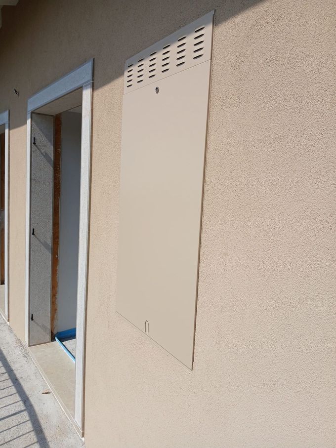 Pannello metallico color marrone chiaro sopra una finestra su una parete in stucco marrone chiaro testurizzato; un'altra finestra è visibile sulla sinistra.