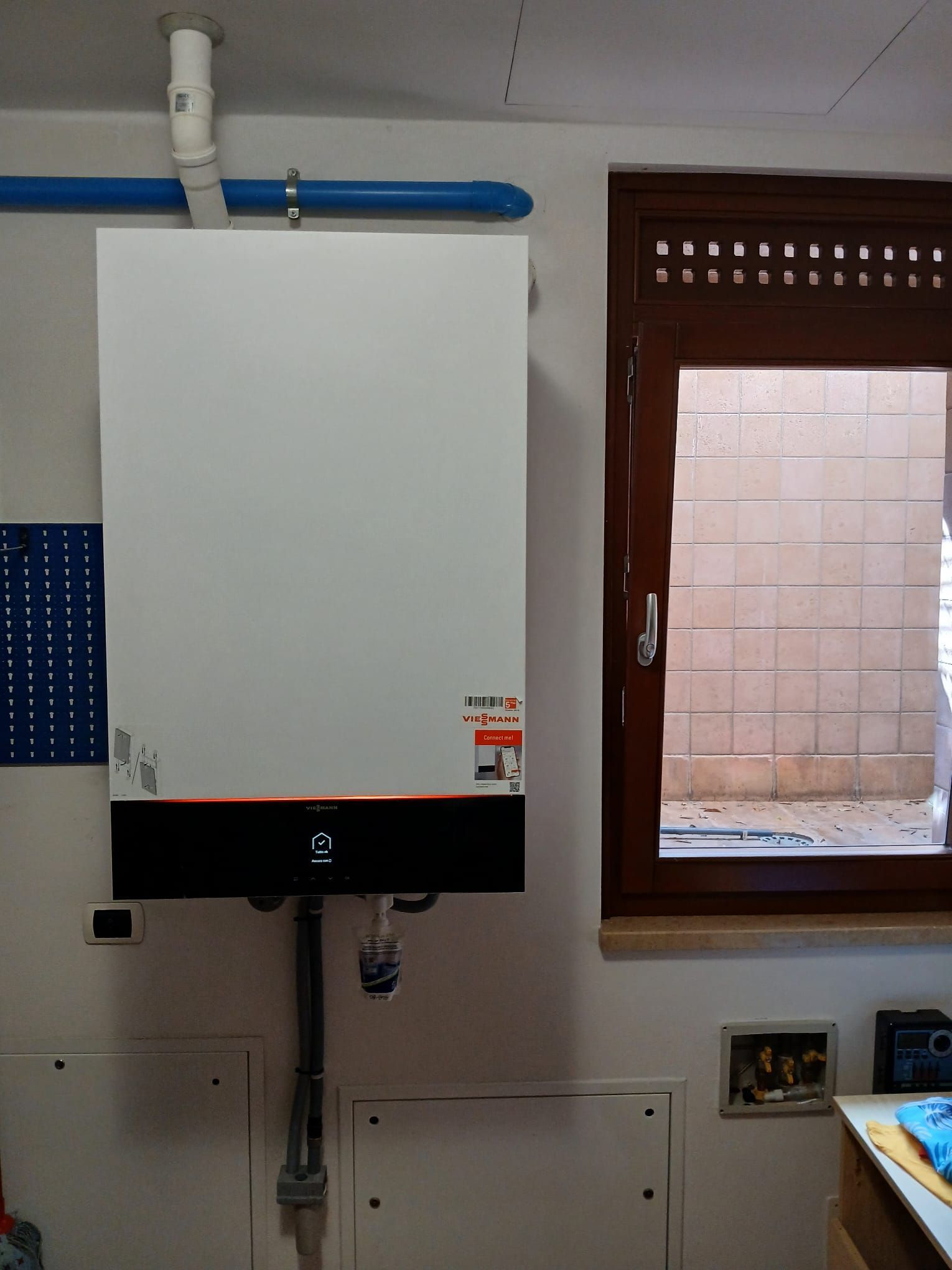 Caldaia bianca a parete con tubo blu, accanto a una finestra e in un ambiente simile a un garage.