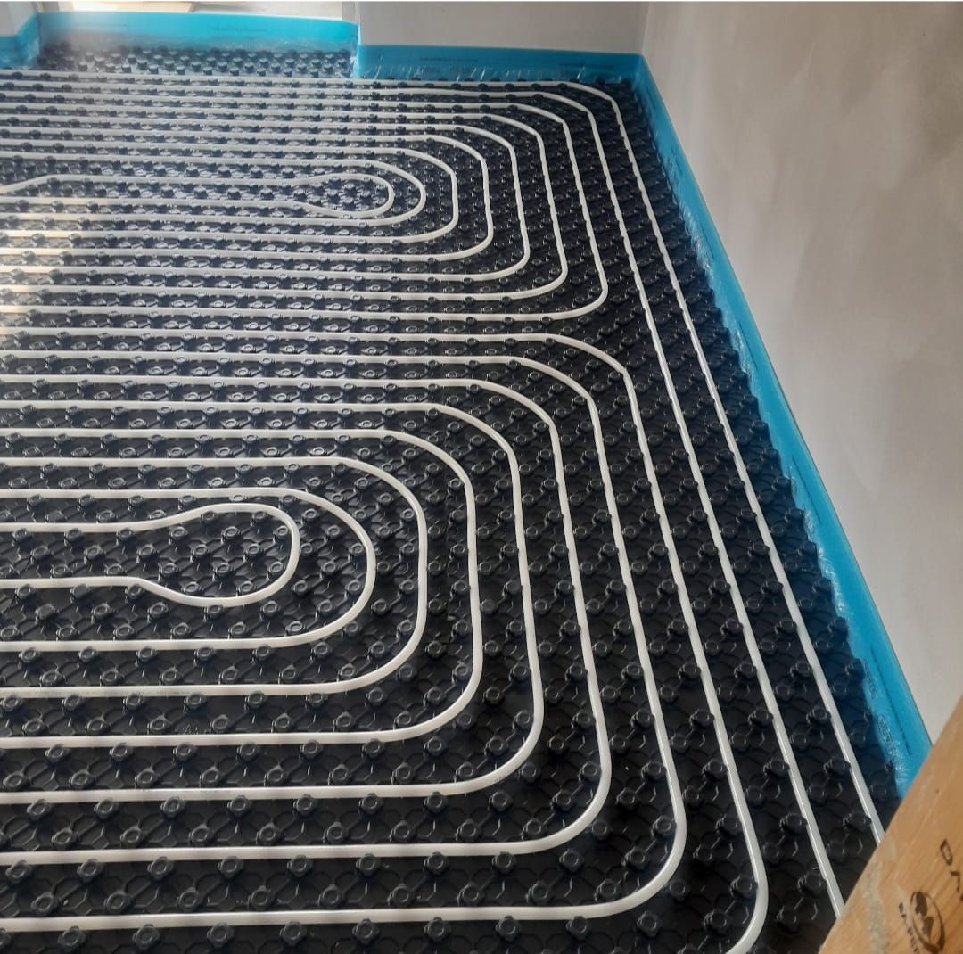Sistema di riscaldamento a pavimento nero con tubi bianchi posati su un isolamento nero, contro una parete nastrata di blu.