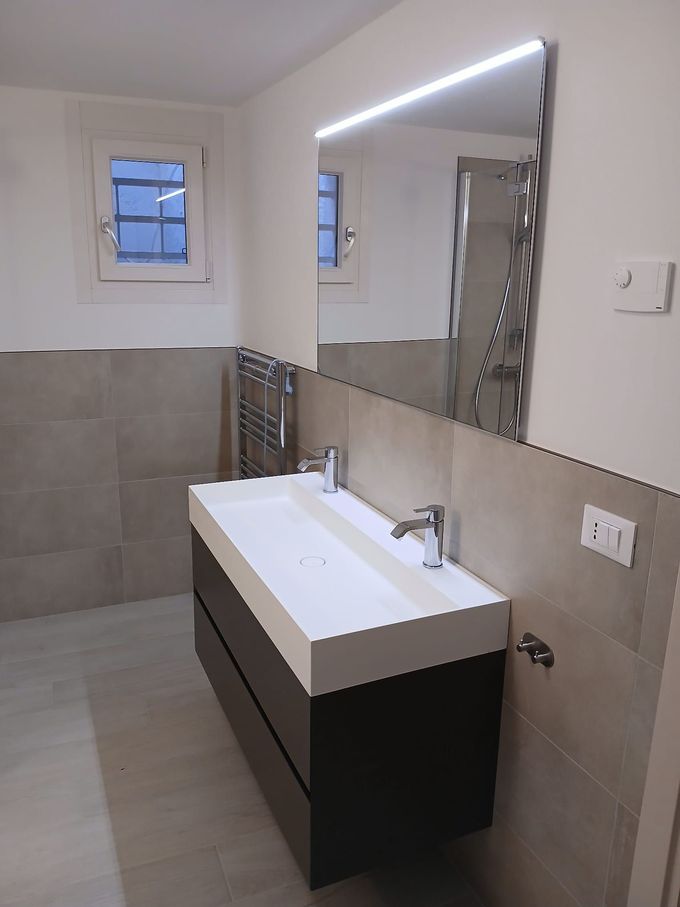Bagno moderno con piastrelle grigie, mobile lavabo scuro, doppio lavandino, grande specchio e piccola finestra.