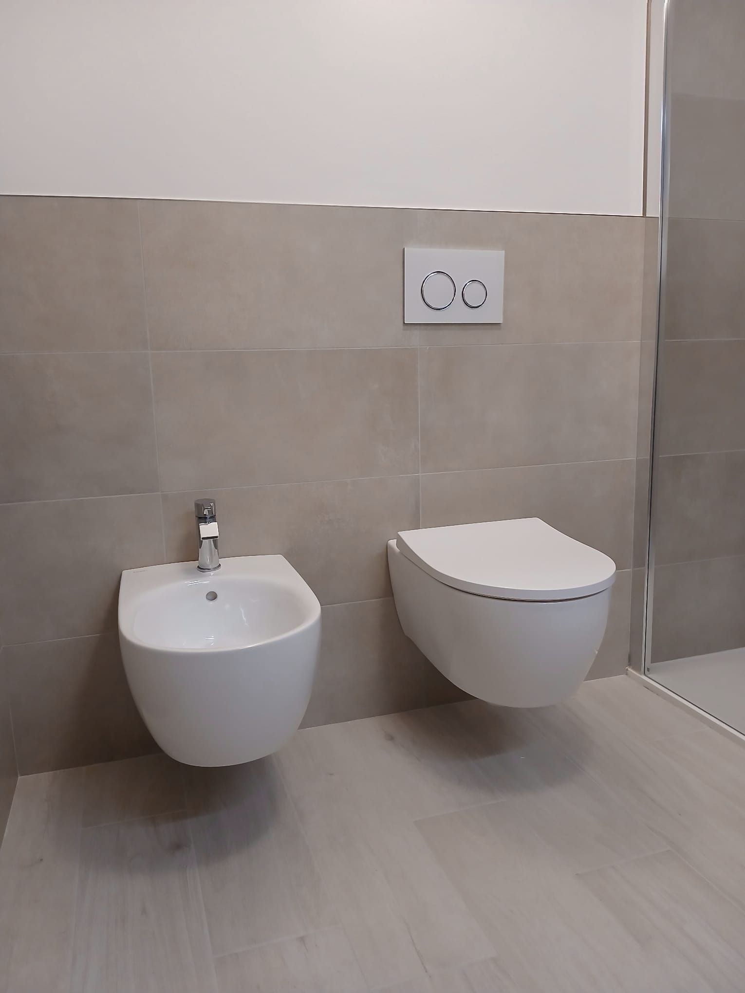 Un bagno con bidet bianco e WC sospeso su pareti e pavimento piastrellati di colore chiaro.