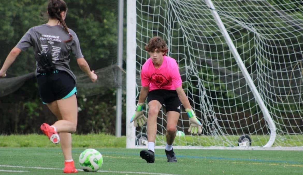 A girl is kicking a soccer ball in front of a goalie.