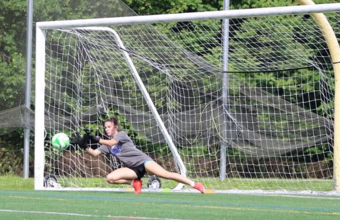 A female soccer goalie is diving to catch a soccer ball.